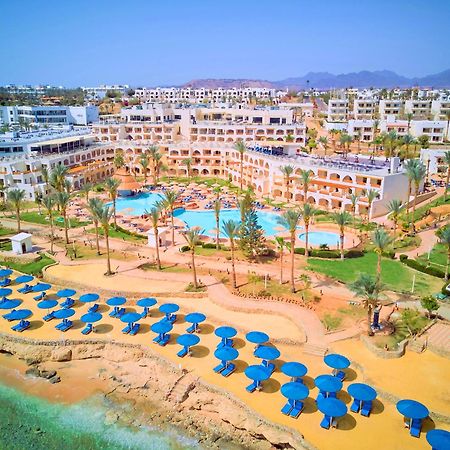 Pickalbatros Royal Grand Sharm - Adults Friendly 16 Years Plus Εξωτερικό φωτογραφία The photo shows a vibrant resort area with several buildings that appear to be accommodations or hotel units. There are multiple swimming pools surrounded by palm trees, creating a tropical atmosphere. In the foreground, there is a beach area with sa