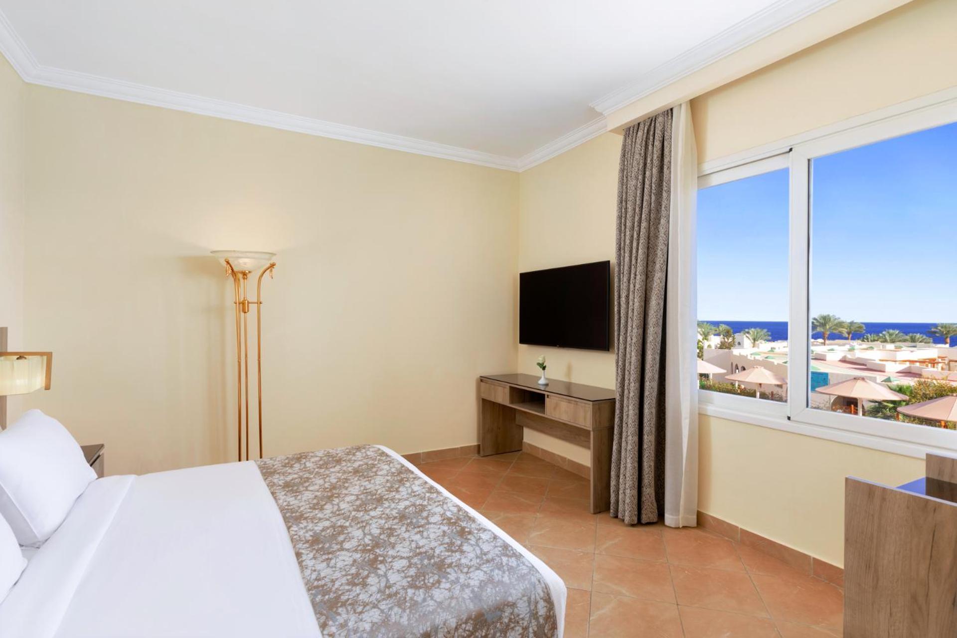 Pickalbatros Royal Grand Sharm - Adults Friendly 16 Years Plus Εξωτερικό φωτογραφία The photo shows a hotel room with a modern and minimalistic design. There is a comfortable bed with white linens and a decorative gray throw at the foot. A large window offers a view of the outdoors, possibly with a glimpse of the sea or beach. A fla