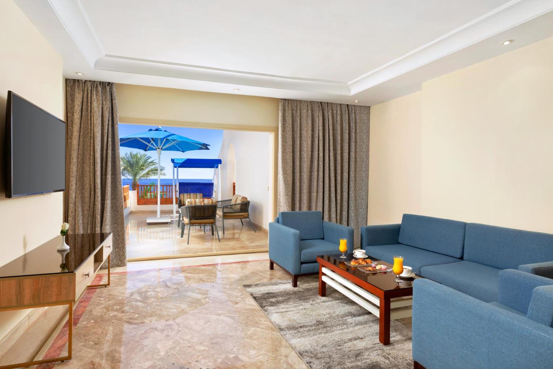 Pickalbatros Royal Grand Sharm - Adults Friendly 16 Years Plus Εξωτερικό φωτογραφία The photo shows a spacious living area with modern decor. There are two blue sofas arranged around a coffee table that holds some decorative items and candles. A flat-screen TV is mounted on the wall across from the seating area. To the left, there a