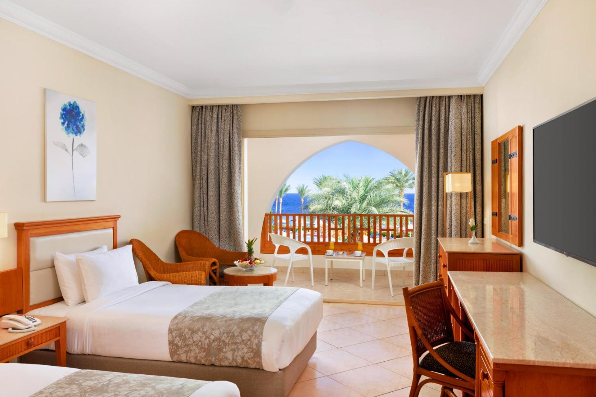 Pickalbatros Royal Grand Sharm - Adults Friendly 16 Years Plus Εξωτερικό φωτογραφία The photo shows a hotel room featuring two beds with light bedding. There is a wooden desk and chair, and a comfortable chair is placed nearby. The room has large windows that open to a balcony with a view of palm trees and a blue sky. The decor is r