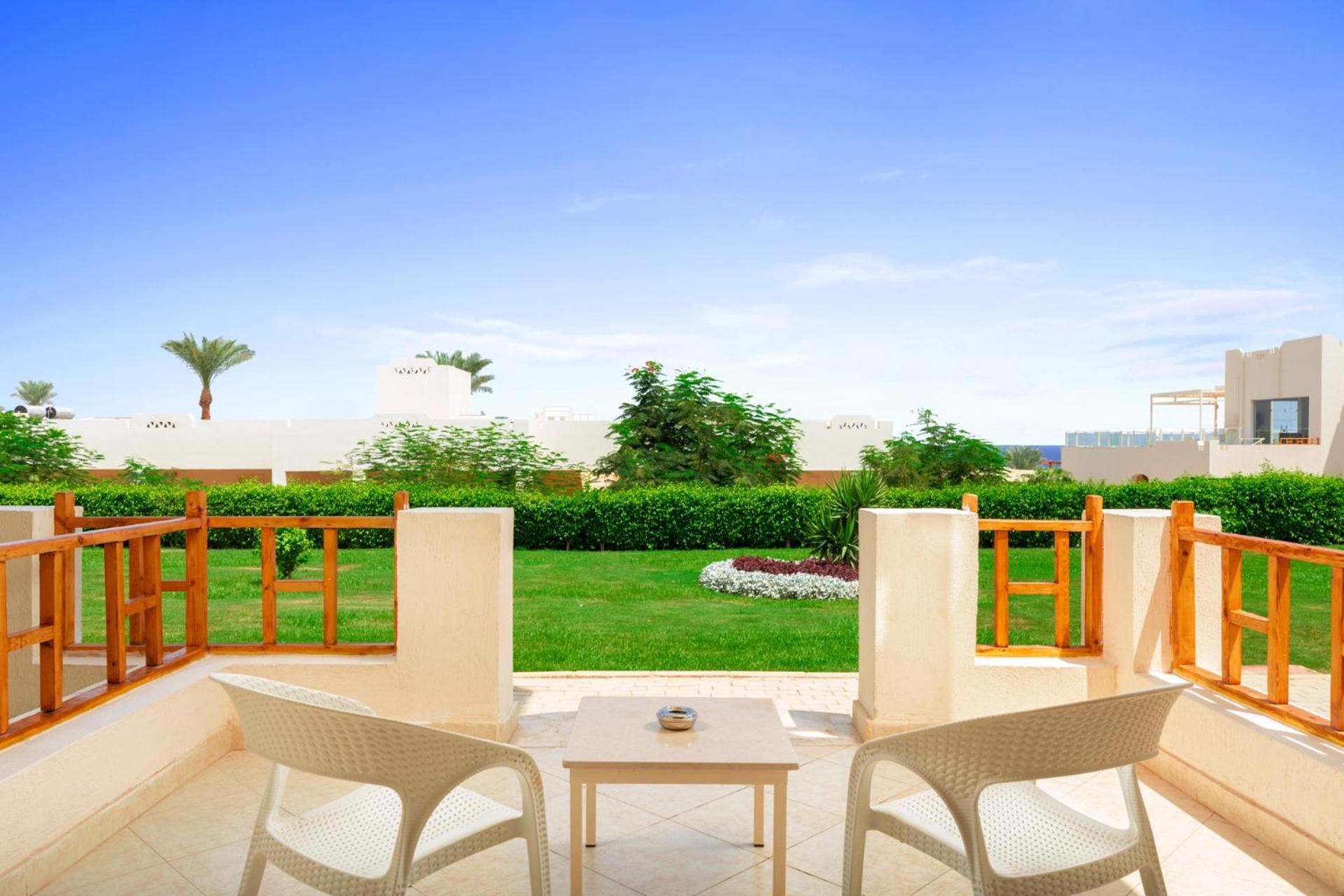 Pickalbatros Royal Grand Sharm - Adults Friendly 16 Years Plus Εξωτερικό φωτογραφία The photo depicts a serene outdoor balcony or patio area. It features two white chairs and a small table, set against a backdrop of lush green grass and garden plants. In the distance, there are some palm trees and clear blue skies, suggesting a warm