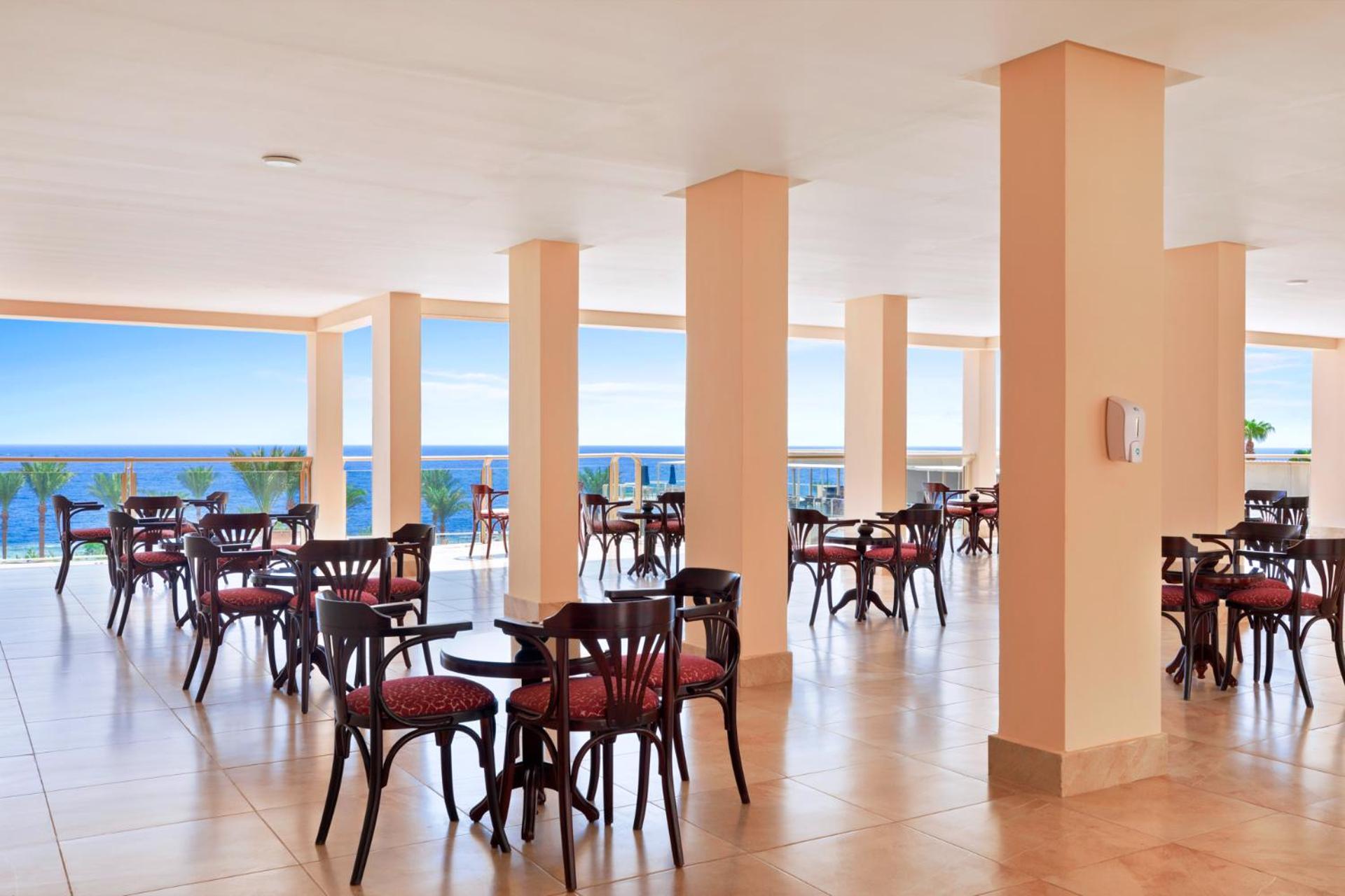 Pickalbatros Royal Grand Sharm - Adults Friendly 16 Years Plus Εξωτερικό φωτογραφία The image shows a dining area or café with several round wooden tables and chairs, featuring red upholstery. The space is well-lit, with large windows that allow a view of the ocean in the background, contributing to a bright and open atmosphere. The