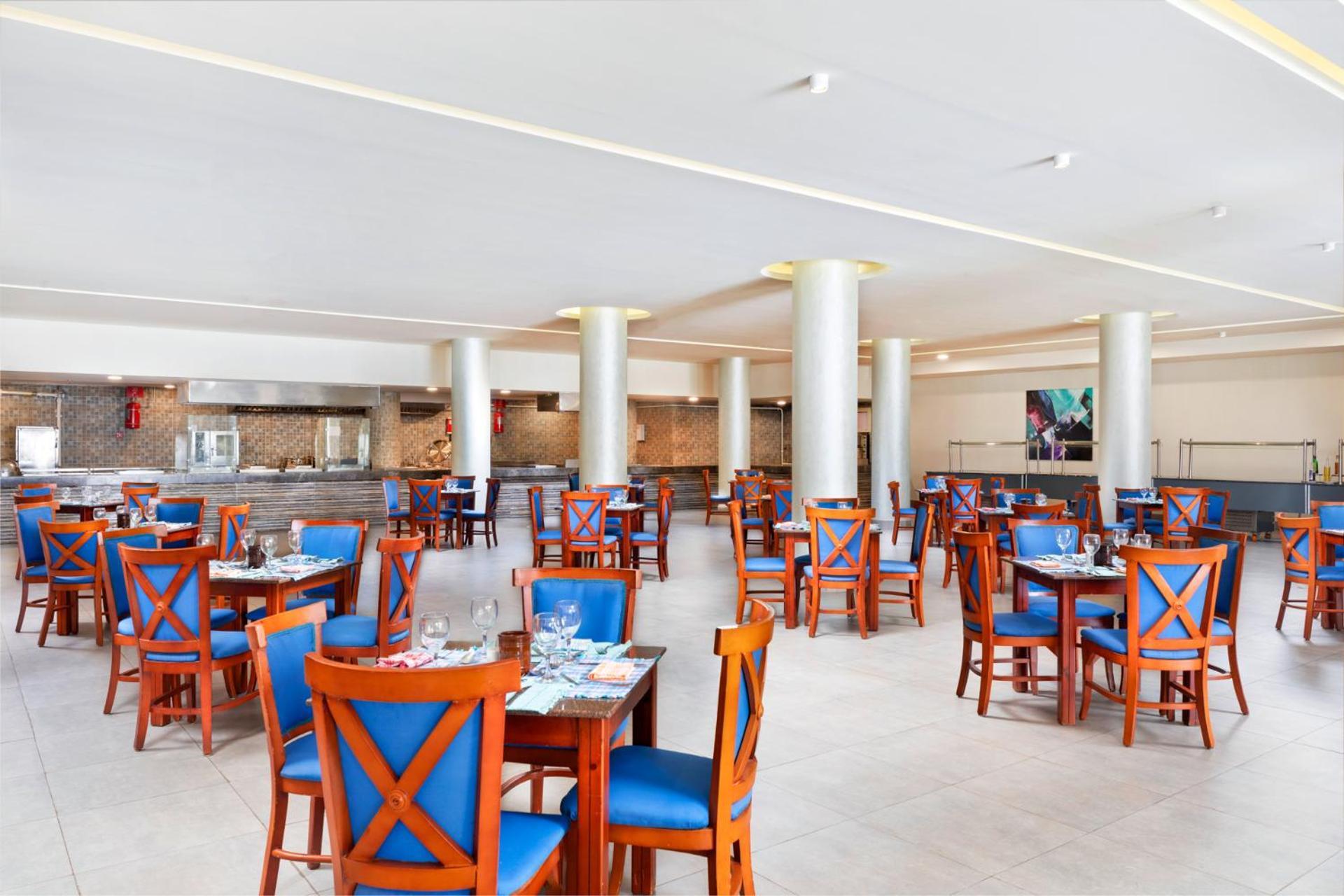 Pickalbatros Royal Grand Sharm - Adults Friendly 16 Years Plus Εξωτερικό φωτογραφία The photo depicts a spacious dining area in a restaurant. The room features several wooden tables, each with blue chairs. The chairs have a unique design with prominent X-shaped backs. The flooring is light-colored tile, and the walls are painted in 
