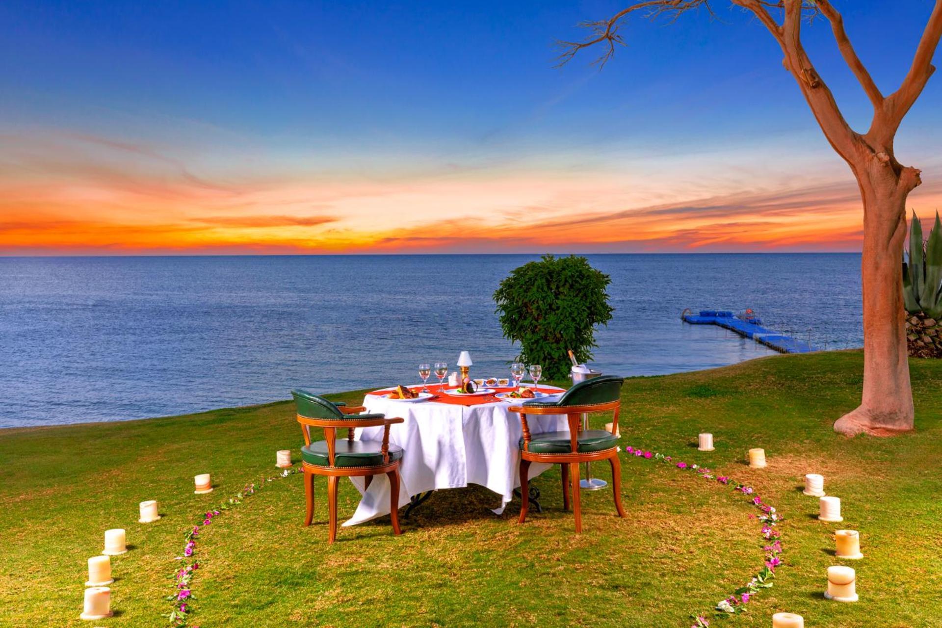 Pickalbatros Royal Grand Sharm - Adults Friendly 16 Years Plus Εξωτερικό φωτογραφία The photo depicts a romantic outdoor dining setup by the seaside. In the foreground, there is a round table elegantly set with a white tablecloth, plates, and glassware, creating a cozy atmosphere. Surrounding the table are green chairs. A circle of 