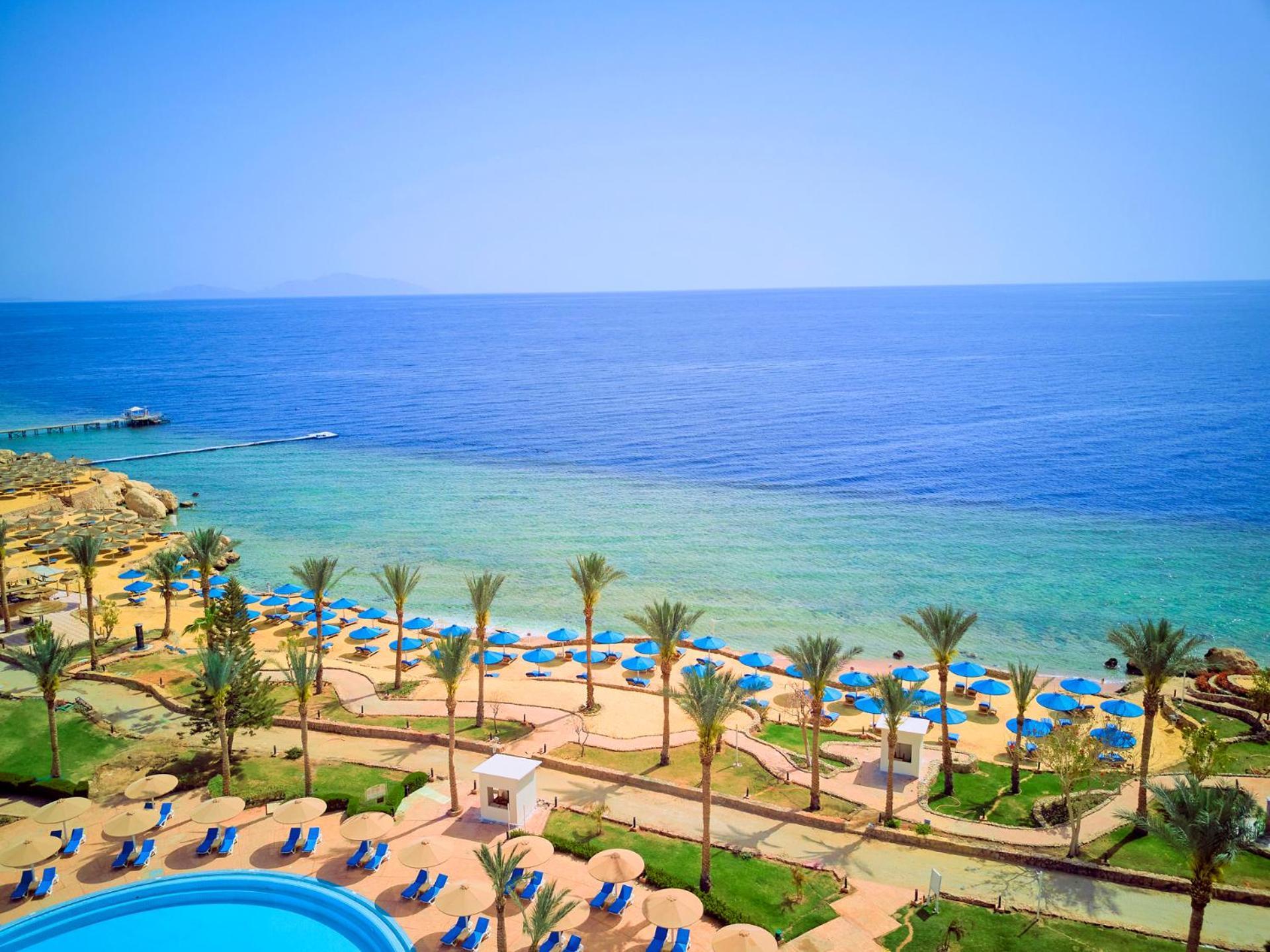 Pickalbatros Royal Grand Sharm - Adults Friendly 16 Years Plus Εξωτερικό φωτογραφία The photo displays a beautiful coastal scene featuring a clear blue sea that transitions into green tones near the shore. In the foreground, there are sandy beaches lined with palm trees and a variety of beach umbrellas. There is also a swimming pool