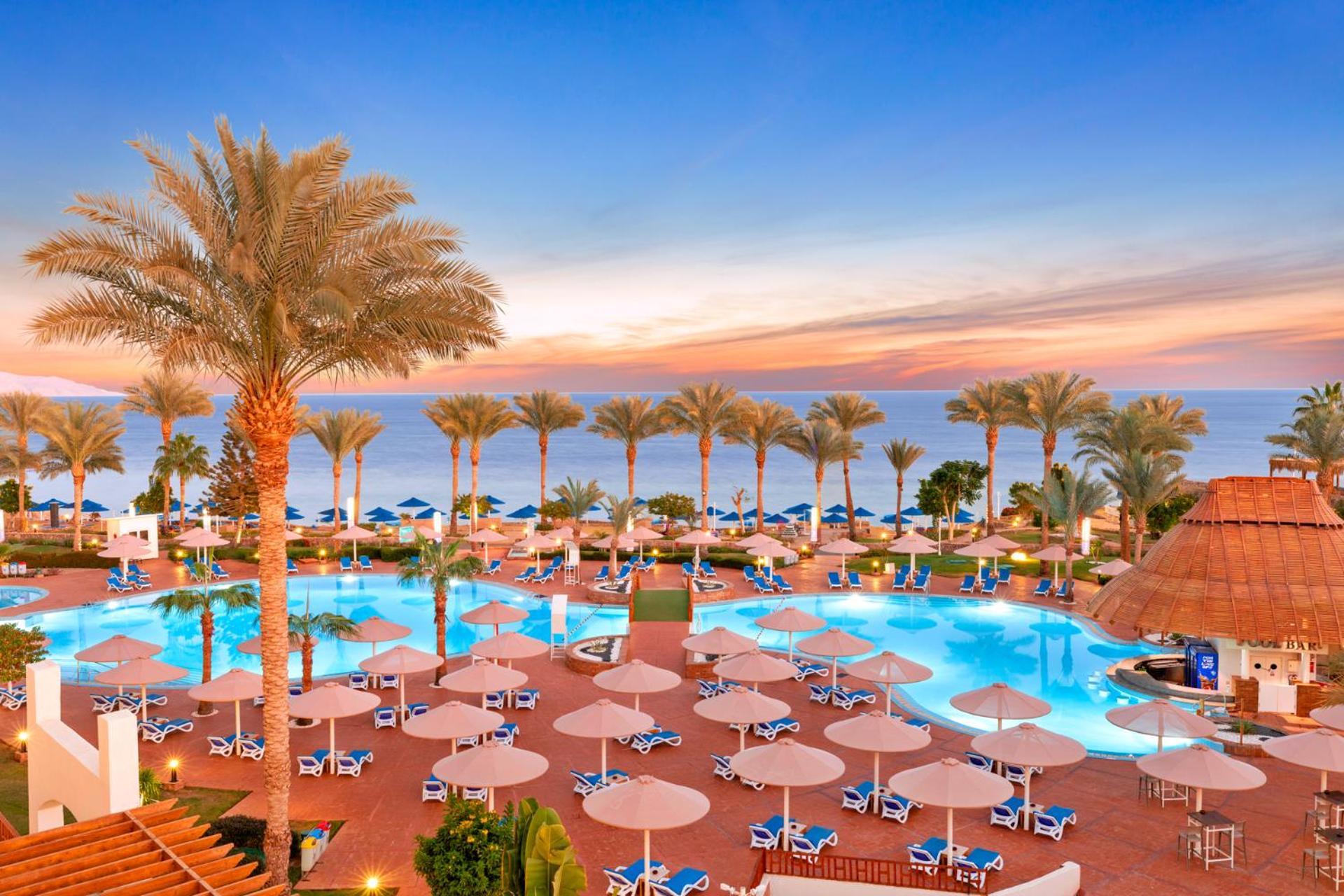 Pickalbatros Royal Grand Sharm - Adults Friendly 16 Years Plus Εξωτερικό φωτογραφία The image shows a beautiful resort setting by the beach. There is a large swimming pool at the center, surrounded by lounge chairs and umbrellas. Palm trees line the area, giving it a tropical vibe. In the background, you can see the ocean and a colo