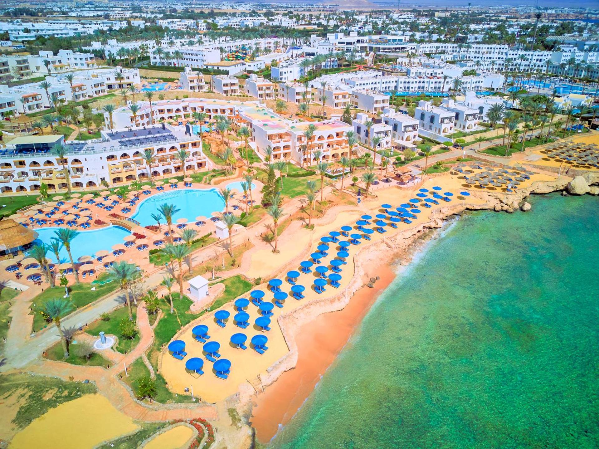 Pickalbatros Royal Grand Sharm - Adults Friendly 16 Years Plus Εξωτερικό φωτογραφία The image shows an aerial view of a resort area by the beach. There is a swimming pool in the center, surrounded by palm trees and sun loungers. The sandy beach features numerous umbrellas arranged in rows for shade. In the background, there are seve
