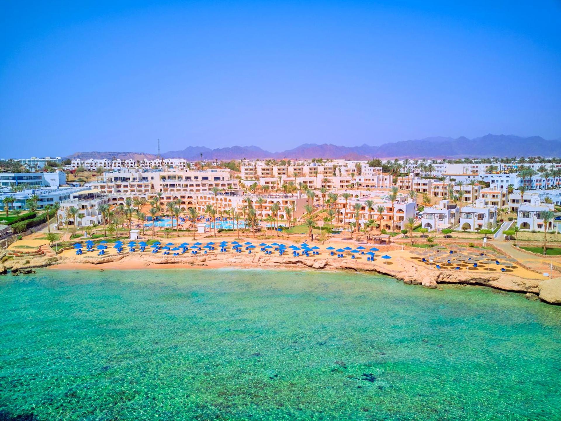 Pickalbatros Royal Grand Sharm - Adults Friendly 16 Years Plus Εξωτερικό φωτογραφία The photo depicts a picturesque beachfront resort. It shows a clear turquoise sea with a sandy shore lined with beach umbrellas and sun loungers. Behind the beach, there are several white buildings of the resort, surrounded by lush greenery. In the b