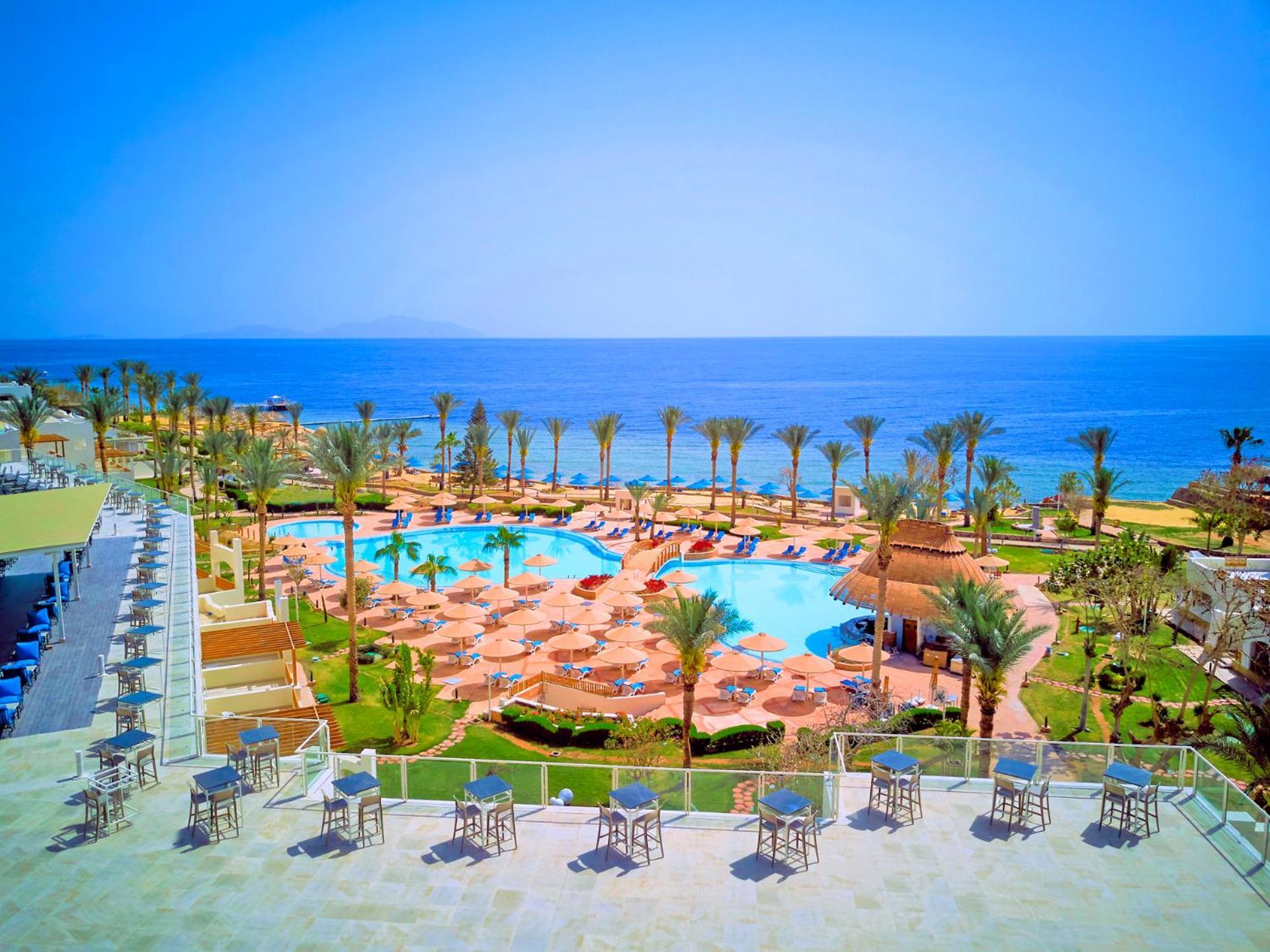 Pickalbatros Royal Grand Sharm - Adults Friendly 16 Years Plus Εξωτερικό φωτογραφία The photo shows a beautiful resort scene with a spacious swimming pool at the center, surrounded by sun loungers and palm trees. There are areas for relaxation with tables and chairs positioned around the pool. In the background, the vibrant blue oce