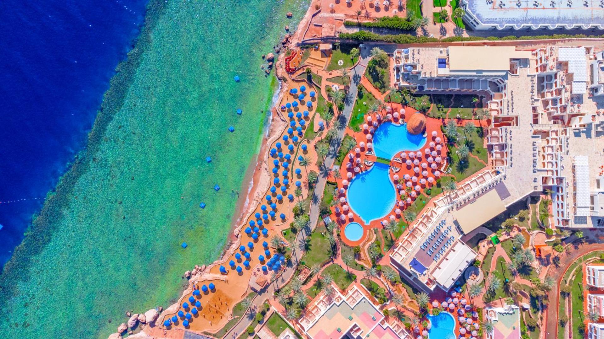 Pickalbatros Royal Grand Sharm - Adults Friendly 16 Years Plus Εξωτερικό φωτογραφία The photo showcases an aerial view of a resort area by the sea. It features a turquoise body of water alongside a sandy beach. There are several umbrellas set up on the beach and lounging areas. A large pool is visible, characterized by its unique sh