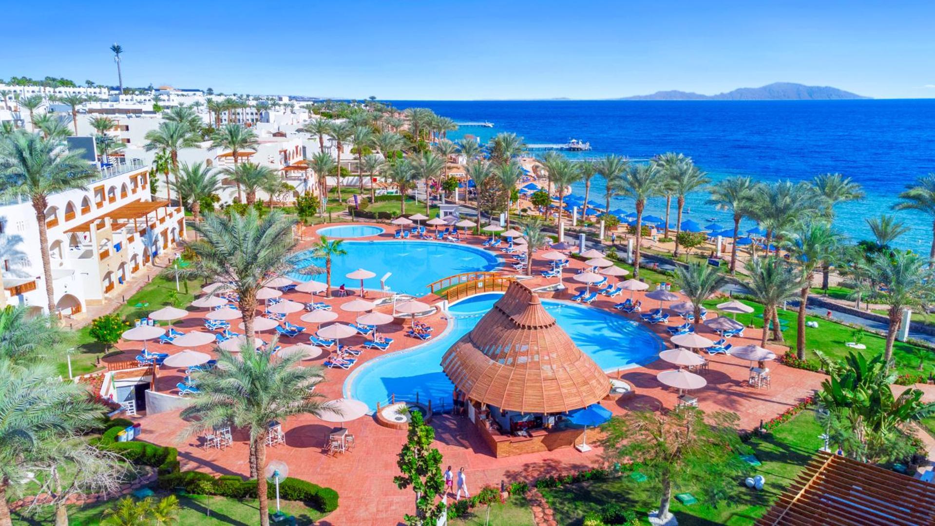 Pickalbatros Royal Grand Sharm - Adults Friendly 16 Years Plus Εξωτερικό φωτογραφία The image depicts a vibrant resort scene overlooking a clear blue sea. In the foreground, there are multiple swimming pools surrounded by sun loungers and umbrellas. A thatched-roof gazebo is situated near the center of the pool area. Palms and lush 