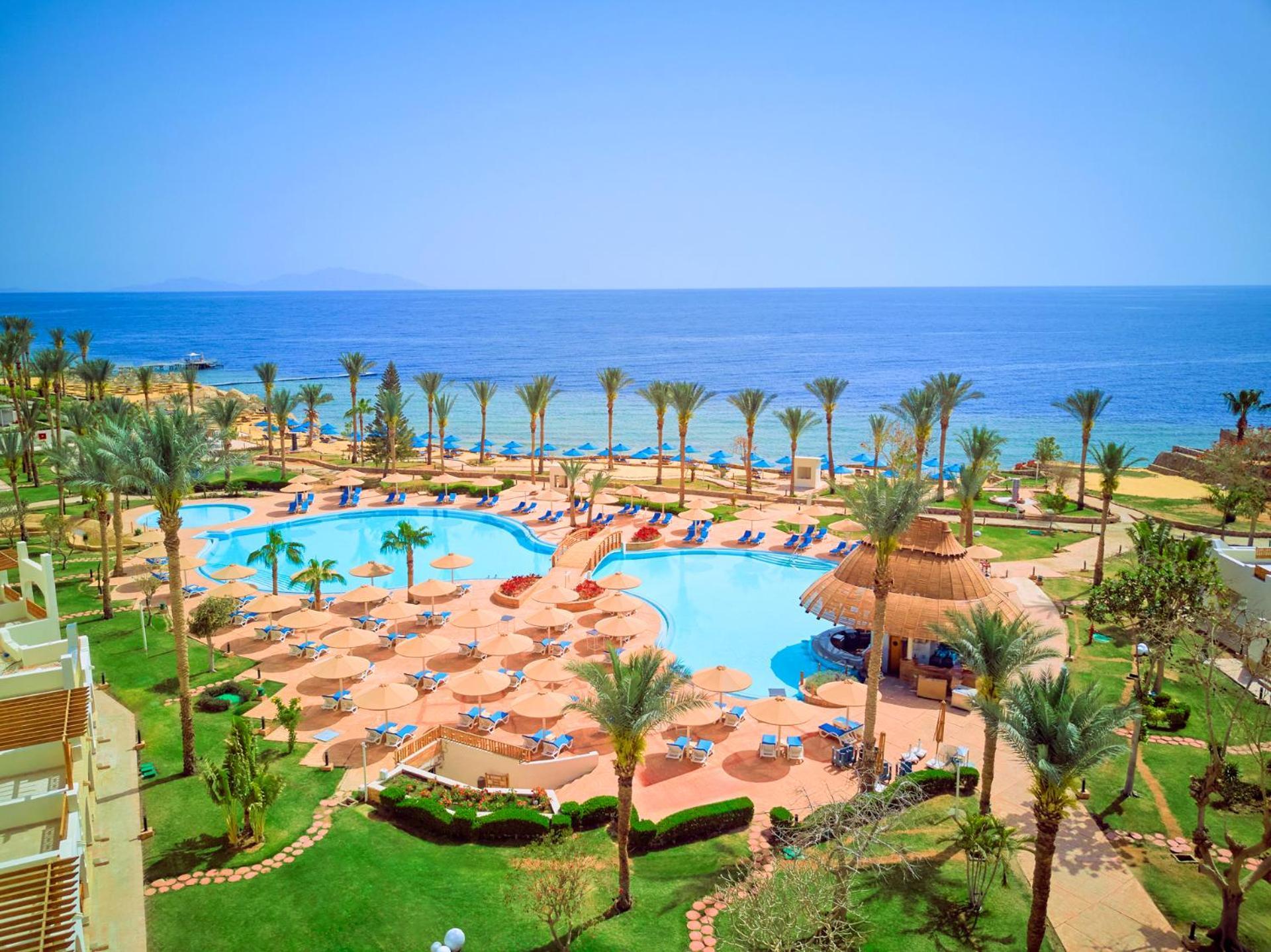 Pickalbatros Royal Grand Sharm - Adults Friendly 16 Years Plus Εξωτερικό φωτογραφία The photo depicts a beautiful resort scene by the ocean. In the foreground, there is a large swimming pool surrounded by palm trees and lounge chairs. The pool area features sunbeds and umbrellas, suggesting a relaxing atmosphere for guests. In the b