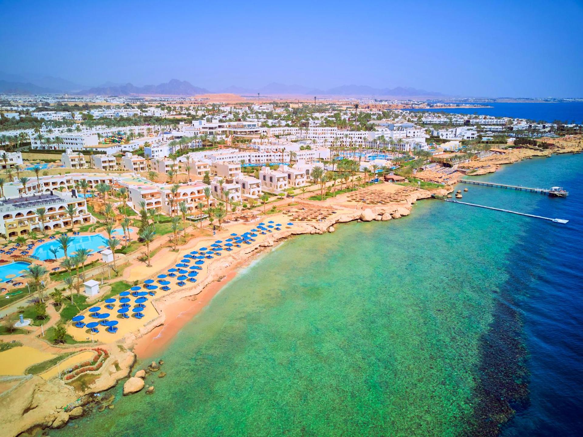 Pickalbatros Royal Grand Sharm - Adults Friendly 16 Years Plus Εξωτερικό φωτογραφία The photo showcases a picturesque coastal scene, likely from a resort area. It features a sandy beach lined with blue umbrellas and lounge chairs, indicating a recreational space for relaxation. The water is clear and exhibits shades of green and blu