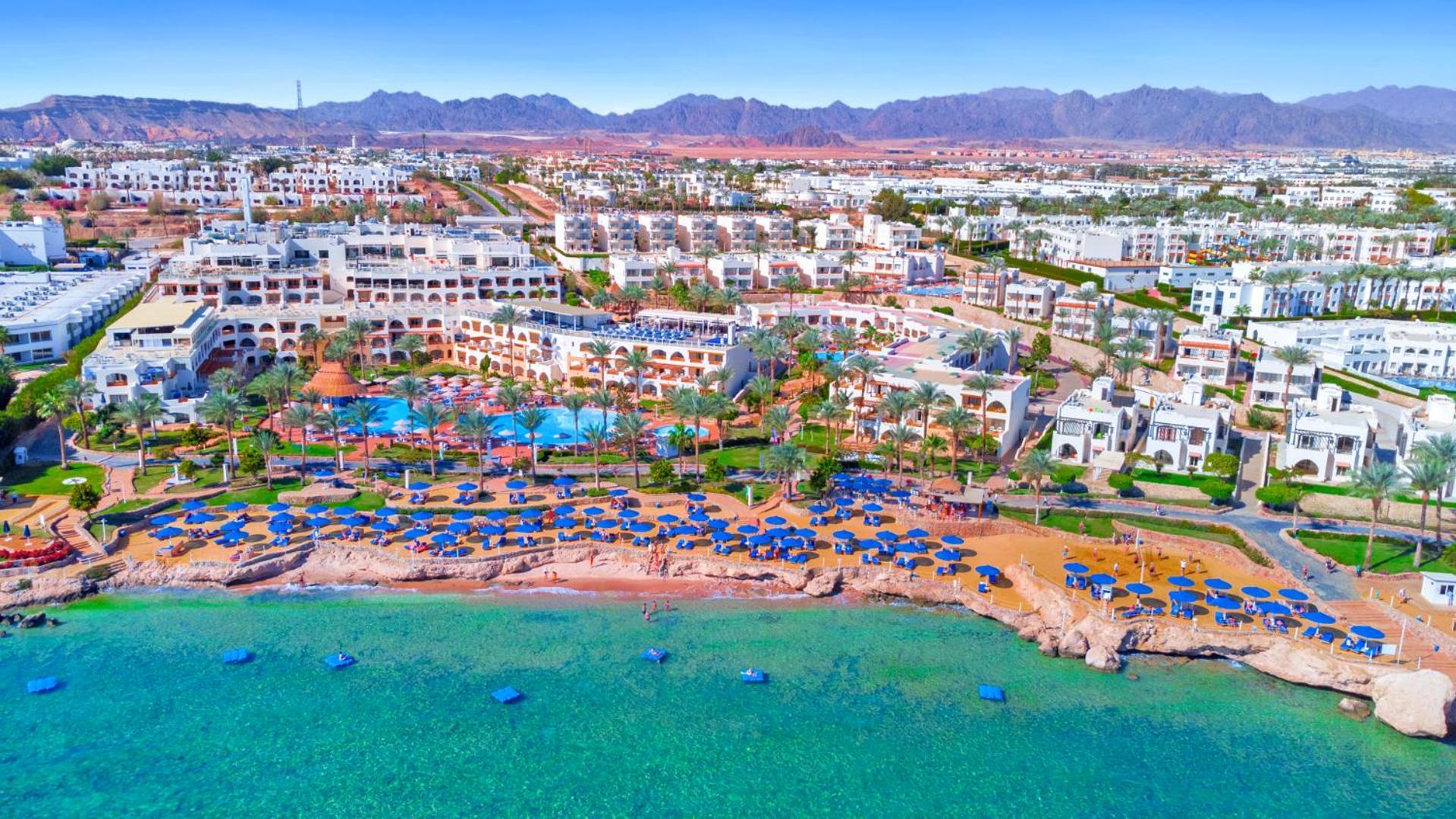 Pickalbatros Royal Grand Sharm - Adults Friendly 16 Years Plus Εξωτερικό φωτογραφία The photo showcases a beautiful coastal resort area. In the foreground, there is a sandy beach lined with blue umbrellas and sun loungers. The vibrant turquoise water gently laps at the shore. Behind the beach, you can see a well-maintained resort wi