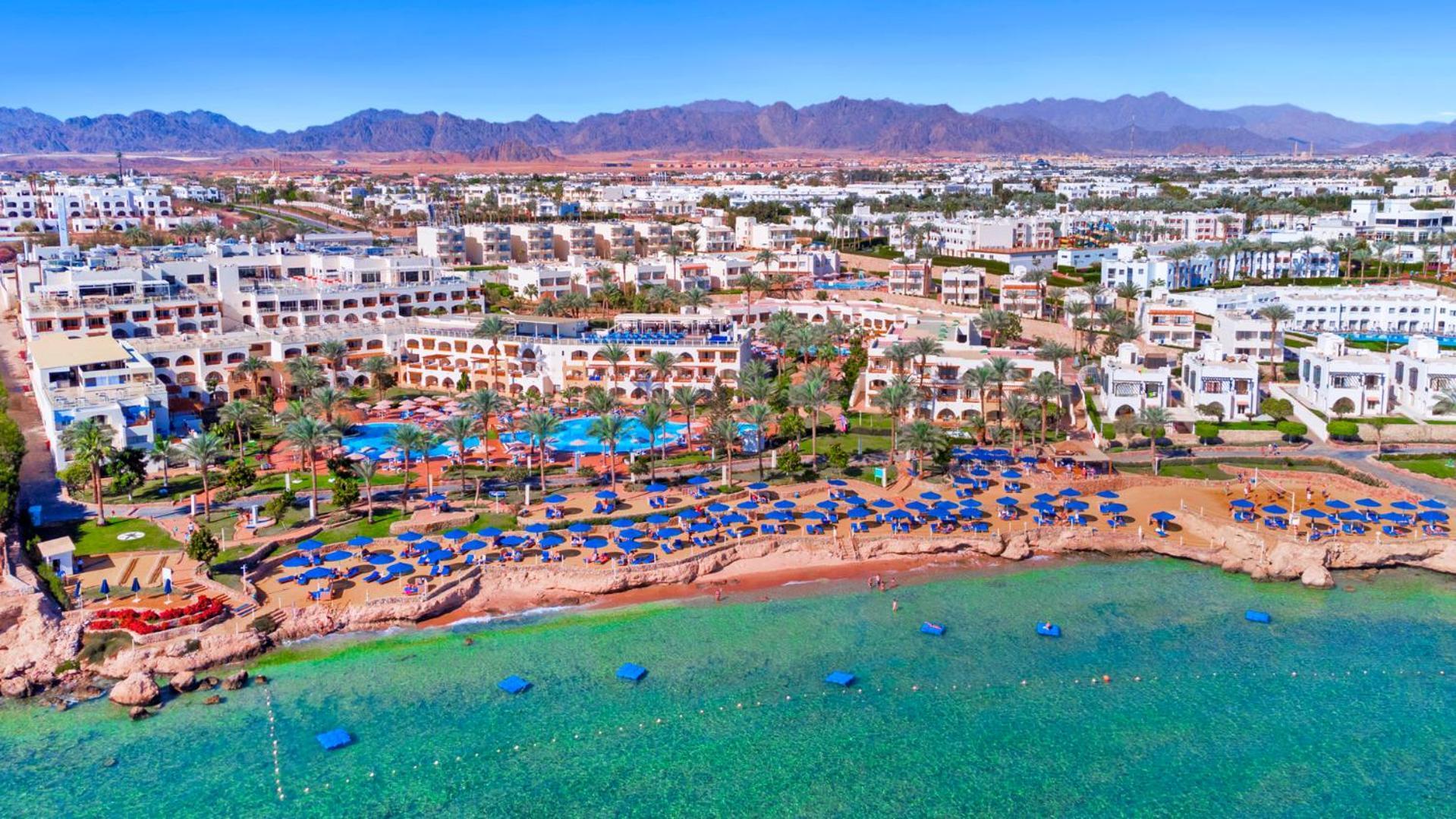 Pickalbatros Royal Grand Sharm - Adults Friendly 16 Years Plus Εξωτερικό φωτογραφία The photo shows an aerial view of a coastal resort area. In the foreground, there is a beach with greenish-blue water, where several blue beach umbrellas and lounges are set up for guests. The shoreline appears sandy, and there are some rocky areas. 