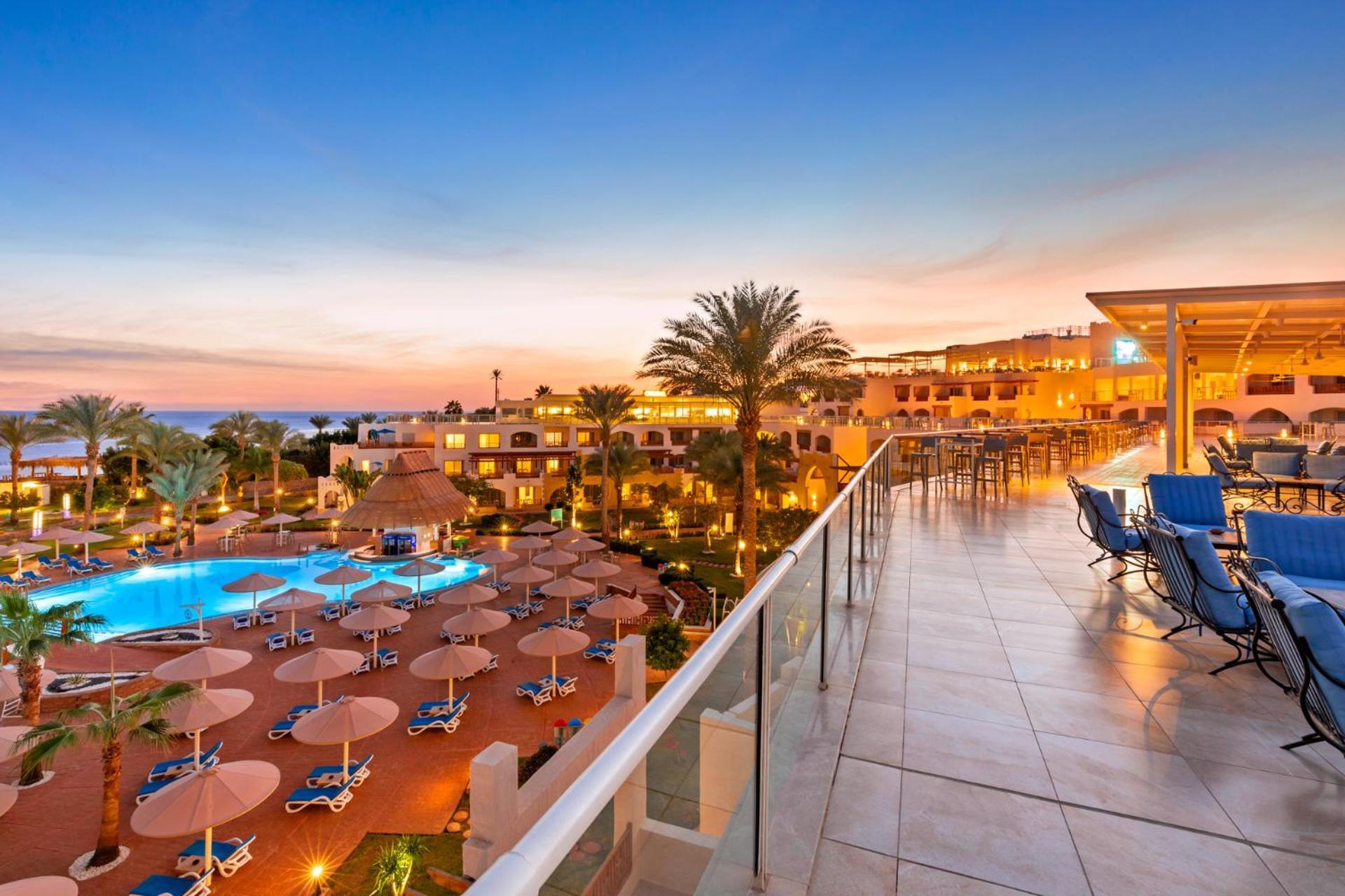 Pickalbatros Royal Grand Sharm - Adults Friendly 16 Years Plus Εξωτερικό φωτογραφία The photo depicts a scenic view of a resort during sunset. In the foreground, there is a balcony with tiled flooring, overlooking the resort. Below, a swimming pool is visible, surrounded by lounge chairs and large umbrellas, indicating a relaxing at