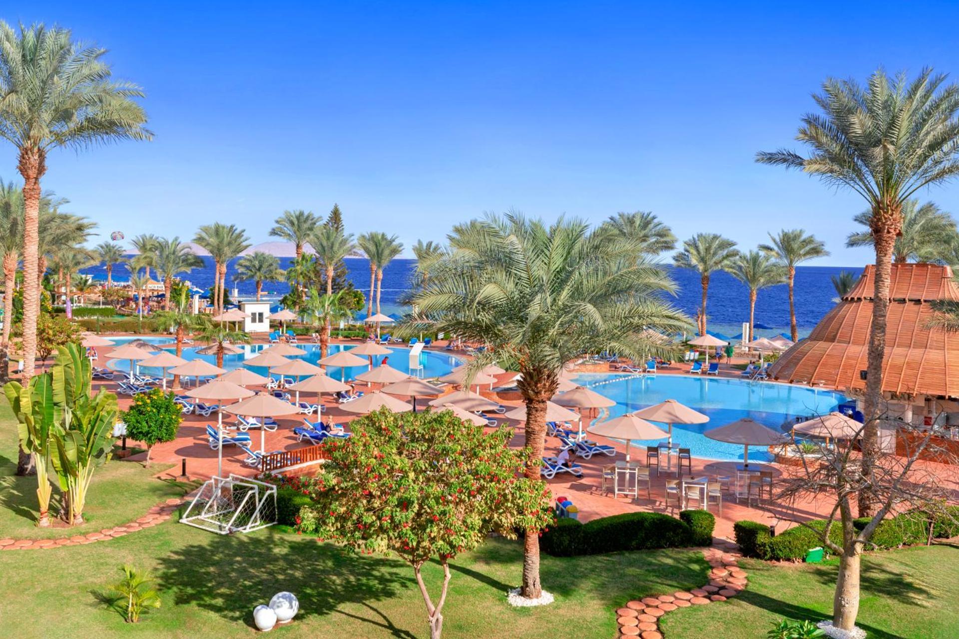 Pickalbatros Royal Grand Sharm - Adults Friendly 16 Years Plus Εξωτερικό φωτογραφία The photo depicts a tropical resort setting by the sea. It features a large swimming pool surrounded by sun loungers and umbrellas. Lush palm trees are scattered throughout the area, providing shade and a picturesque backdrop. In the background, you 