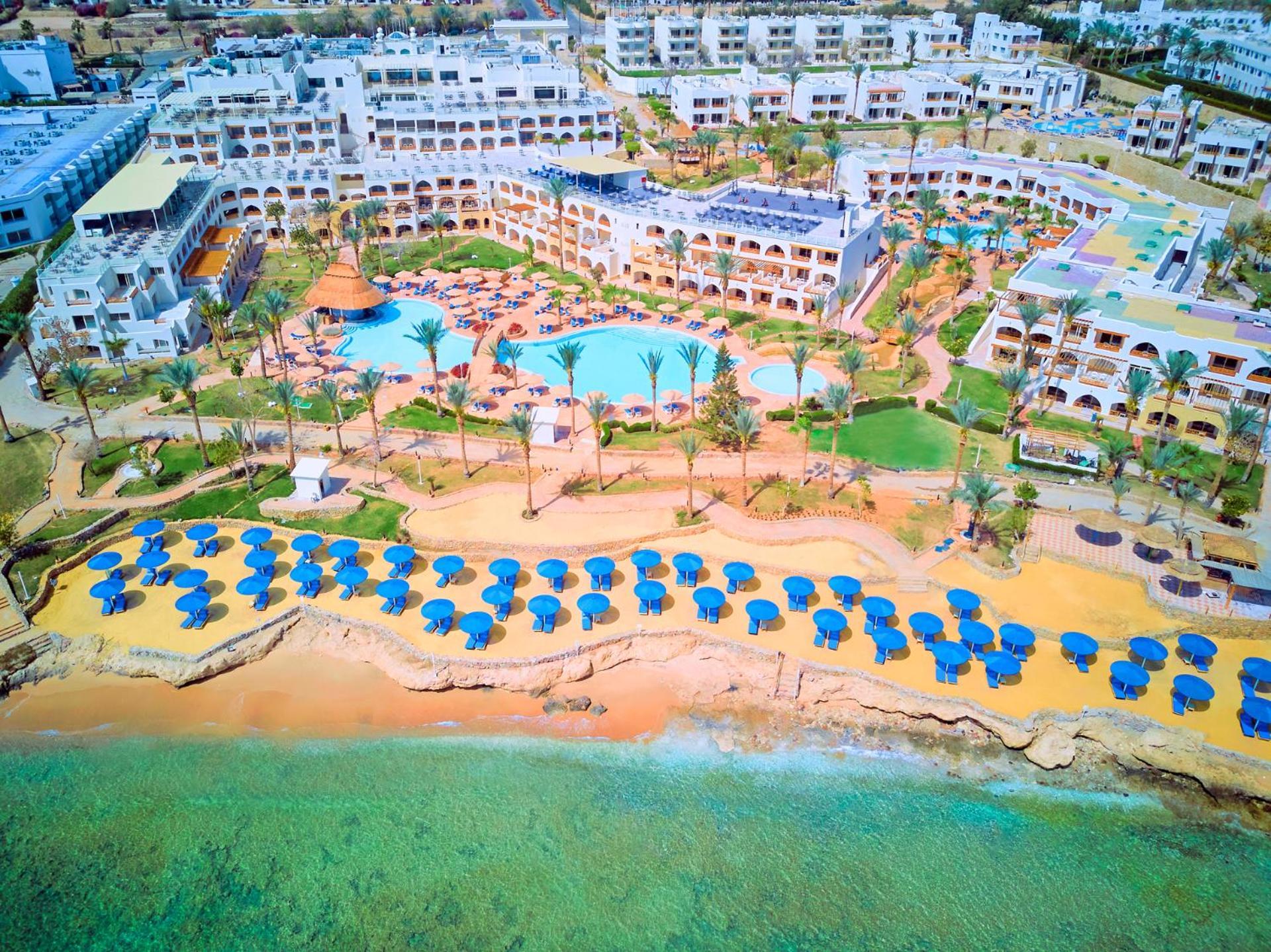 Pickalbatros Royal Grand Sharm - Adults Friendly 16 Years Plus Εξωτερικό φωτογραφία The photo shows a large resort complex situated near a beach. There is a turquoise swimming pool at the center, surrounded by green lawns and palm trees. Sun loungers with blue umbrellas are lined up on the sandy beach, and the beach area is adjacent