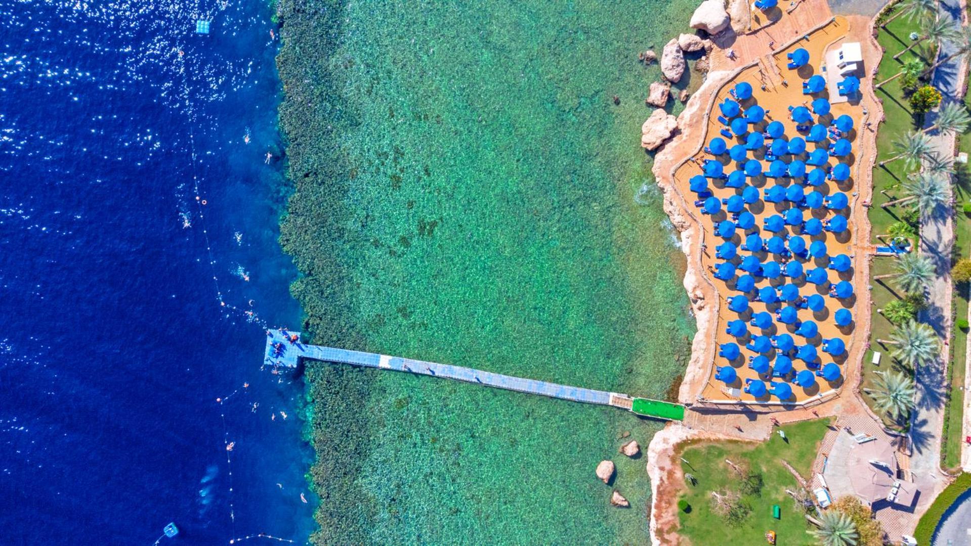 Pickalbatros Royal Grand Sharm - Adults Friendly 16 Years Plus Εξωτερικό φωτογραφία The photo shows an aerial view of a beach area. In the foreground, there is a wooden pier extending into the clear blue water. To the right, there are numerous beach umbrellas with blue canopies arranged neatly, likely indicating a resort or beach cl