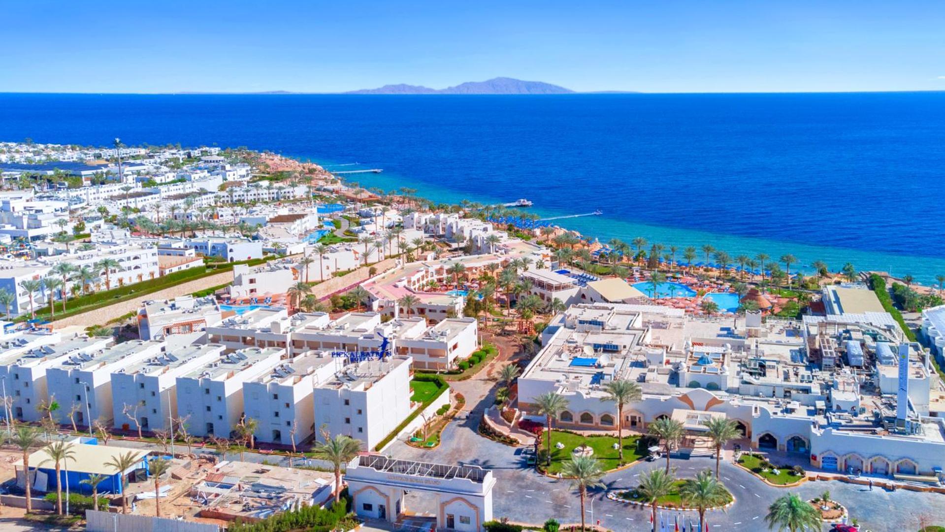 Pickalbatros Royal Grand Sharm - Adults Friendly 16 Years Plus Εξωτερικό φωτογραφία The photo shows a beautiful coastal landscape, featuring a resort area by the sea. There are white buildings, likely hotels or villas, arranged in a sprawling layout near the beach. The water is a vibrant blue, contrasting with the sandy shore and gr