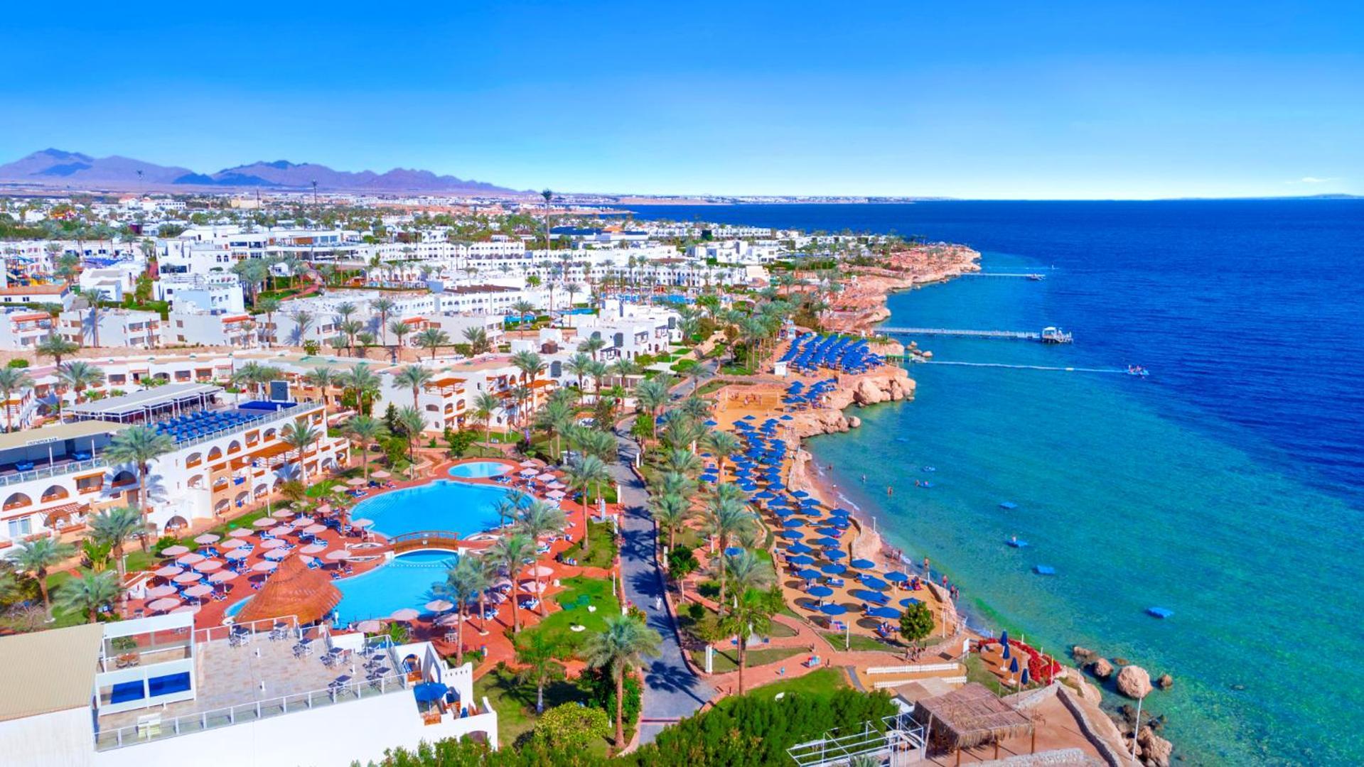 Pickalbatros Royal Grand Sharm - Adults Friendly 16 Years Plus Εξωτερικό φωτογραφία The photo features a picturesque coastal scene, likely from a resort area. In the foreground, there is a large swimming pool surrounded by lounge chairs. Further back, the landscape includes several buildings, possibly hotels or apartments, that over
