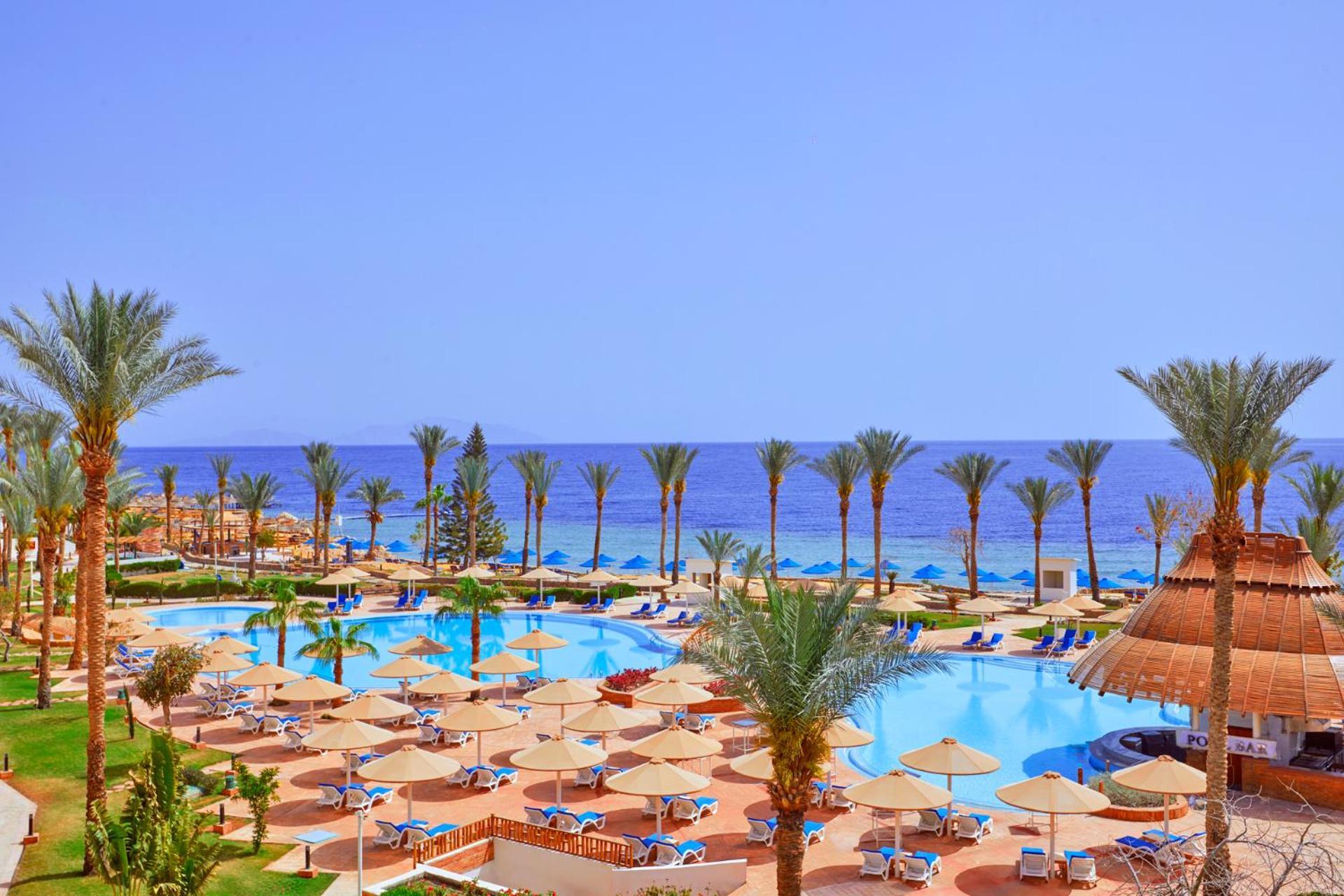 Pickalbatros Royal Grand Sharm - Adults Friendly 16 Years Plus Εξωτερικό φωτογραφία The photo showcases a beautiful resort setting by the sea. It features a large swimming pool surrounded by sunny lounge chairs and umbrellas, perfect for relaxation. The area is adorned with lush palm trees, adding to the tropical vibe. In the backgr