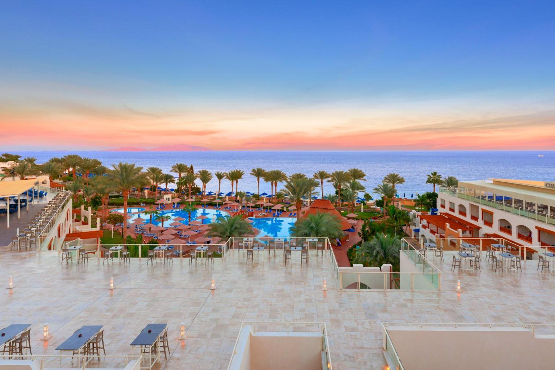Pickalbatros Royal Grand Sharm - Adults Friendly 16 Years Plus Εξωτερικό φωτογραφία The photo depicts a beautiful resort scene at sunset. In the foreground, there is a spacious terrace area with modern furniture. Below, a large swimming pool is surrounded by lush palm trees and lounge chairs, indicating a relaxing atmosphere. The vi