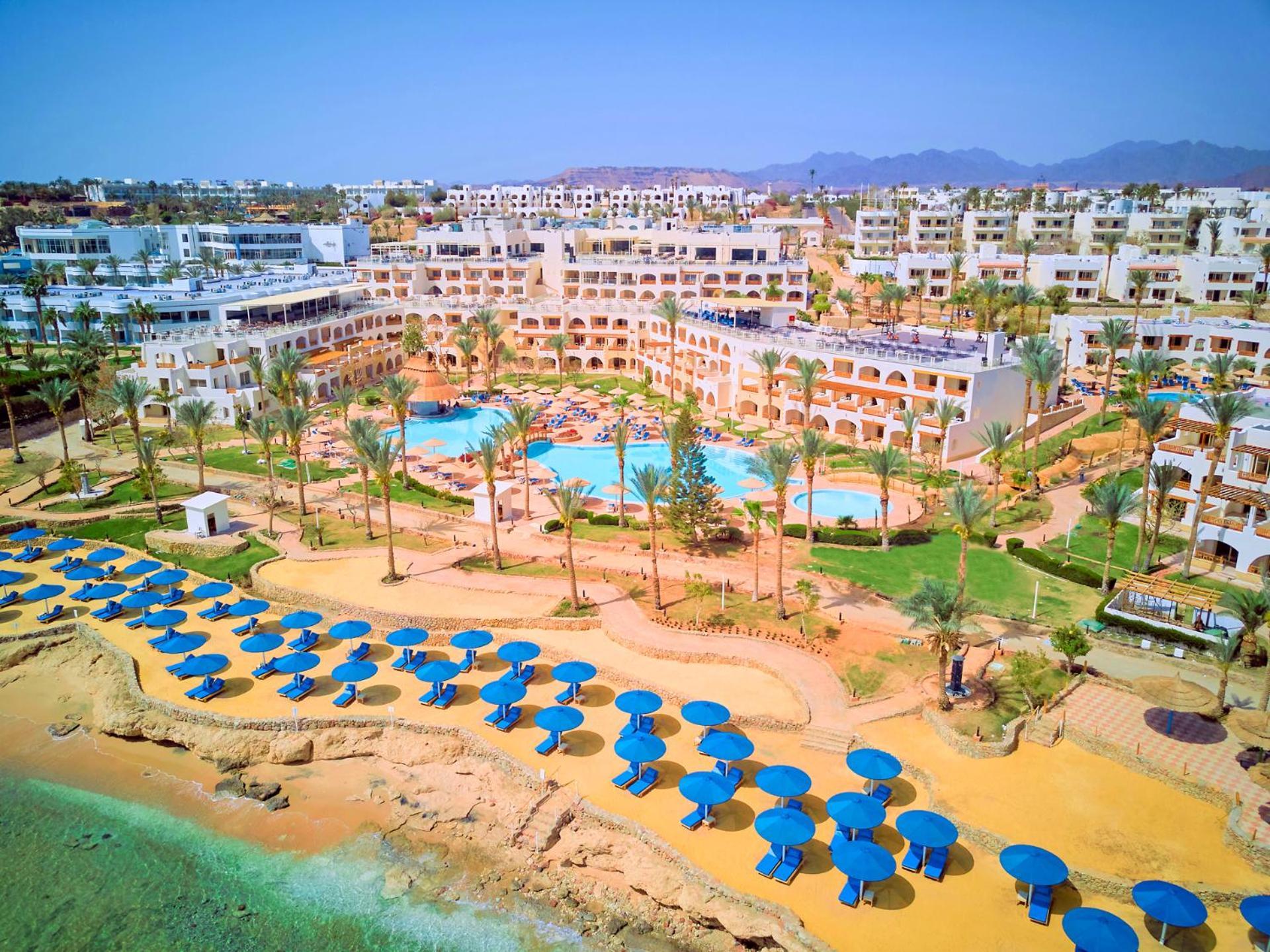 Pickalbatros Royal Grand Sharm - Adults Friendly 16 Years Plus Εξωτερικό φωτογραφία The photo shows a vibrant resort area with several buildings that appear to be accommodations or hotel units. There are multiple swimming pools surrounded by palm trees, creating a tropical atmosphere. In the foreground, there is a beach area with sa
