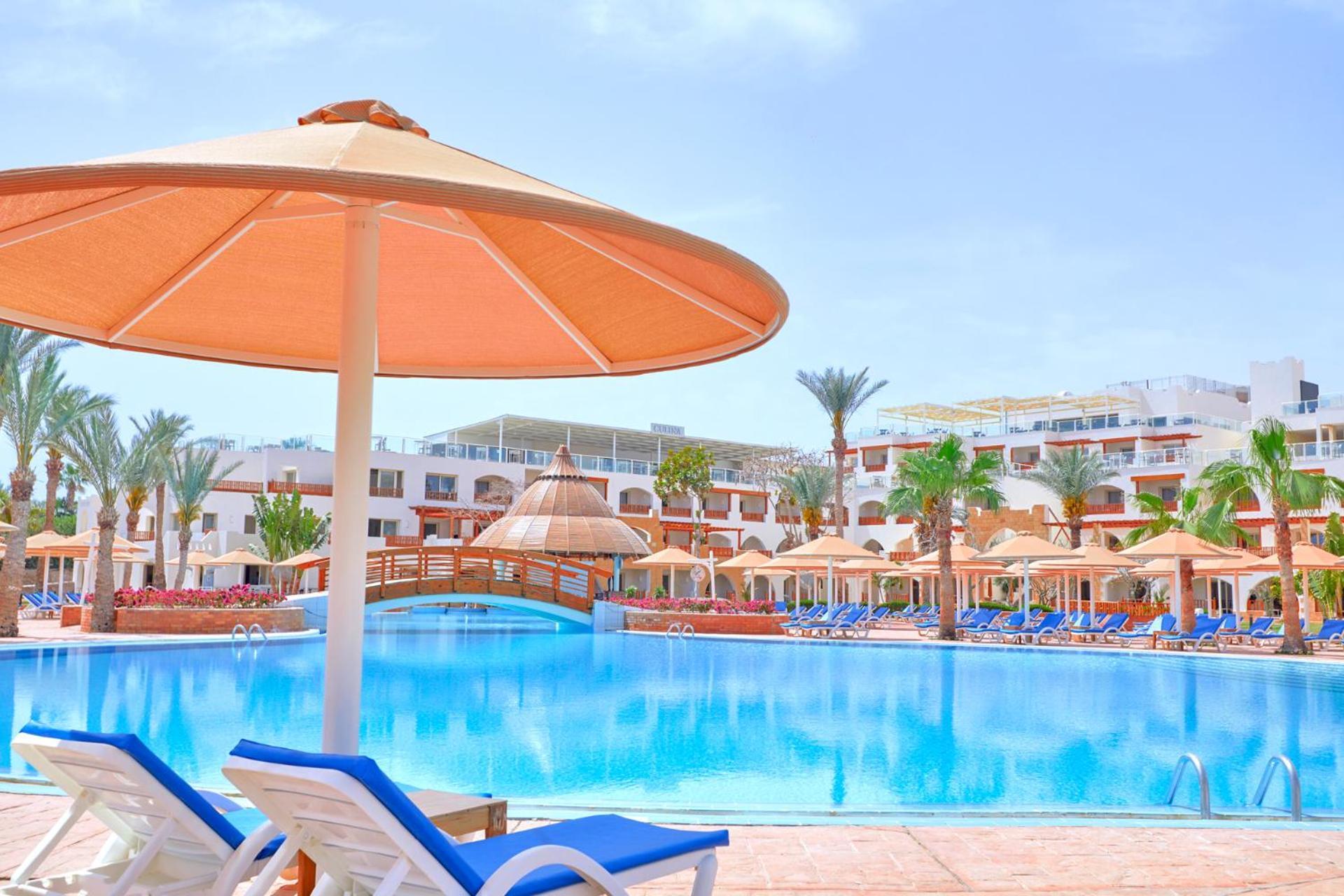 Pickalbatros Royal Grand Sharm - Adults Friendly 16 Years Plus Εξωτερικό φωτογραφία The photo shows a picturesque resort scene featuring a large swimming pool surrounded by lounge chairs and umbrellas. The pool is shimmering under a bright blue sky, and there are several palm trees in the background, adding to the tropical atmospher