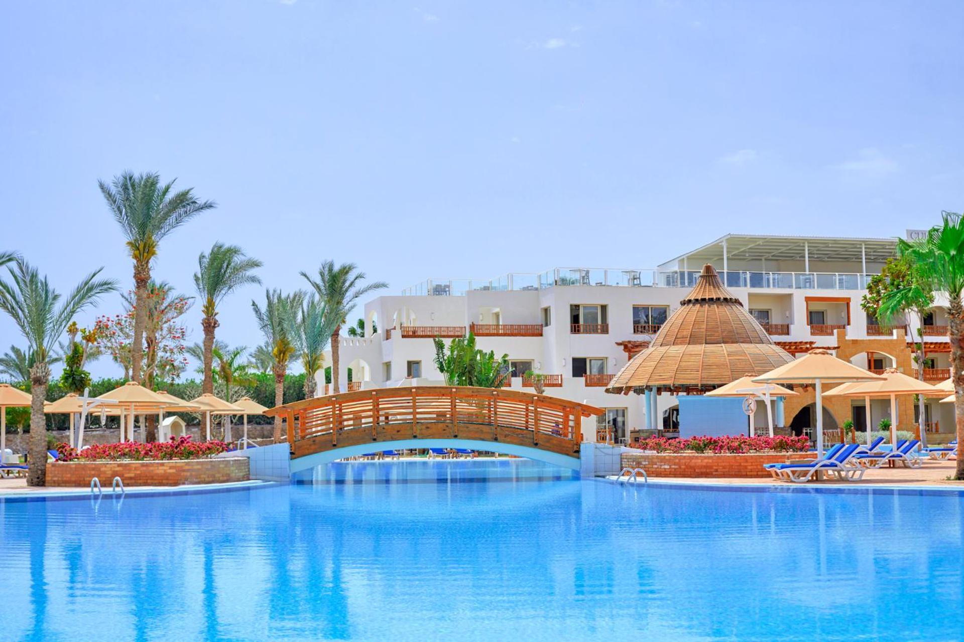 Pickalbatros Royal Grand Sharm - Adults Friendly 16 Years Plus Εξωτερικό φωτογραφία The photo depicts a serene resort setting featuring a large, calm swimming pool, which reflects the clear blue sky. In the foreground, a wooden bridge arches over the pool. Surrounding the pool are several palm trees and tropical foliage, contributin