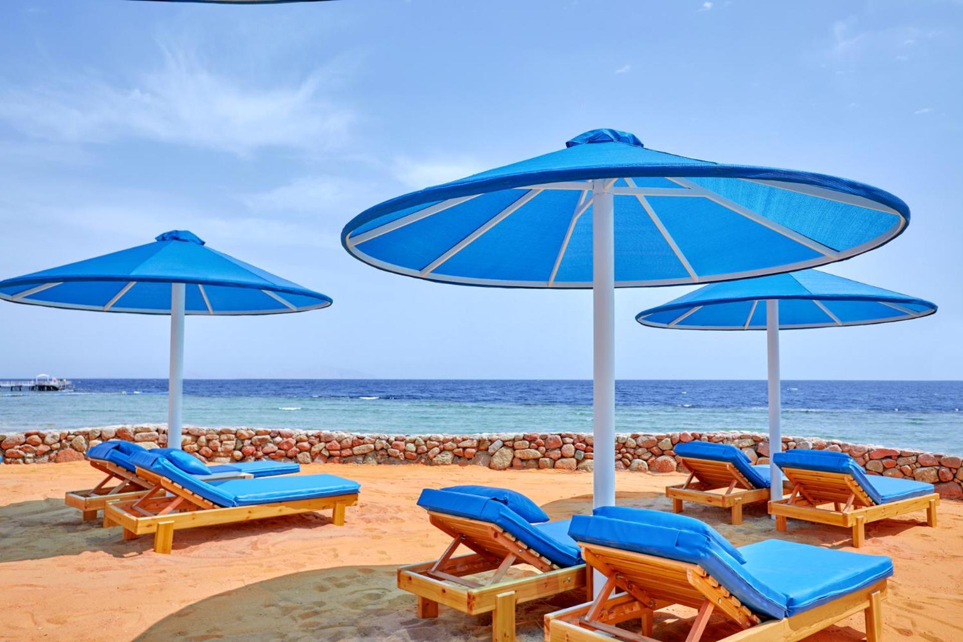 Pickalbatros Royal Grand Sharm - Adults Friendly 16 Years Plus Εξωτερικό φωτογραφία The photo shows a serene beach scene with several blue sun loungers arranged beneath large blue umbrellas. The loungers are positioned on sandy ground, with the ocean visible in the background. The sky is clear, indicating a bright and sunny day, and