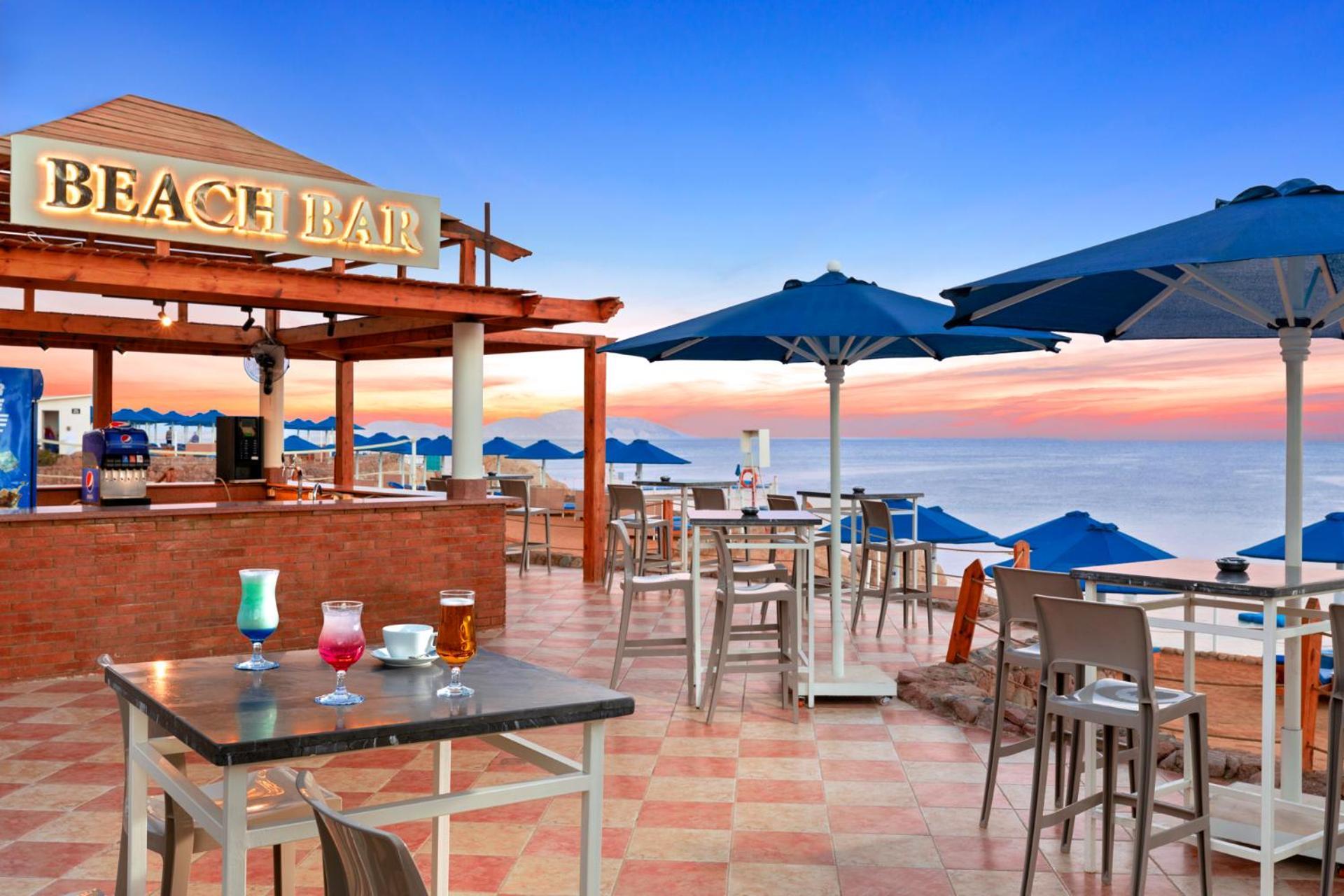 Pickalbatros Royal Grand Sharm - Adults Friendly 16 Years Plus Εξωτερικό φωτογραφία The photo shows a beach bar setting, featuring a wooden structure with the words "BEACH BAR" prominently displayed. There are several tables and chairs arranged on a tiled floor, with some drinks placed on the tables. Blue umbrellas provide shade for
