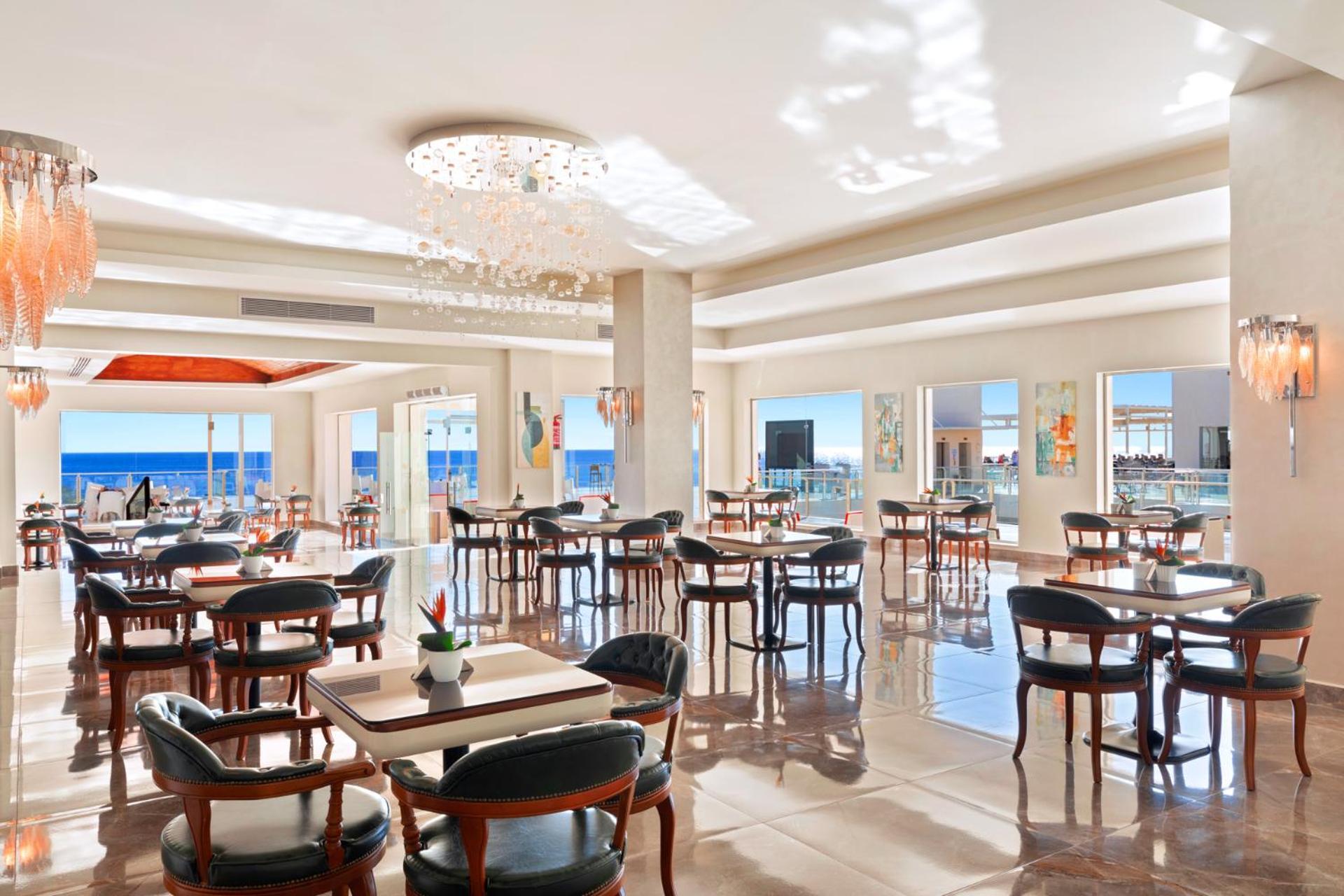 Pickalbatros Royal Grand Sharm - Adults Friendly 16 Years Plus Εξωτερικό φωτογραφία The photo shows a bright and elegant restaurant interior. There are several tables arranged throughout the space, each with chairs around them. The decor features modern lighting, including a chandelier, and large windows that allow natural light to 