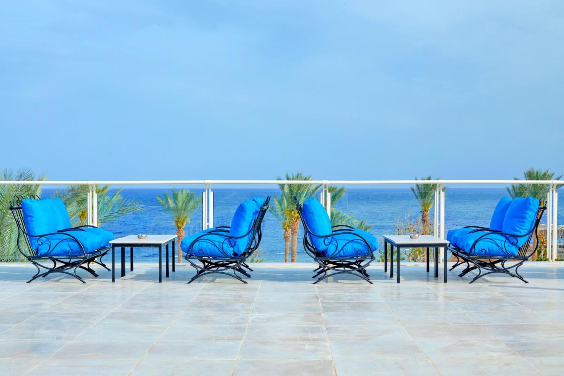 Pickalbatros Royal Grand Sharm - Adults Friendly 16 Years Plus Εξωτερικό φωτογραφία The photo depicts a serene outdoor patio area with four blue lounge chairs arranged around small tables. The setting features a clear view of the ocean with palm trees visible in the background, complementing the tranquil atmosphere. The overall colo