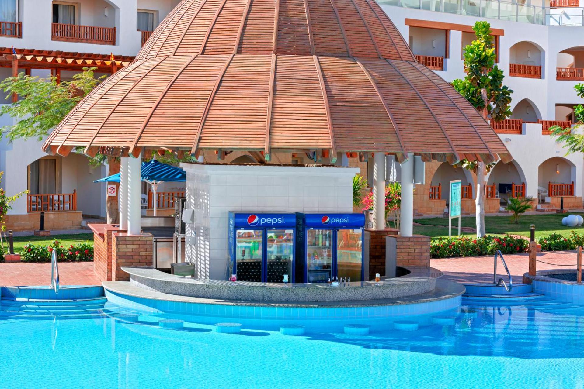 Pickalbatros Royal Grand Sharm - Adults Friendly 16 Years Plus Εξωτερικό φωτογραφία The photo shows a poolside bar with a thatched roof structure, located near a swimming pool. The bar features a counter where drinks are served, and there are vending machines displaying the Pepsi brand. Surrounding the bar, there are neatly landscap
